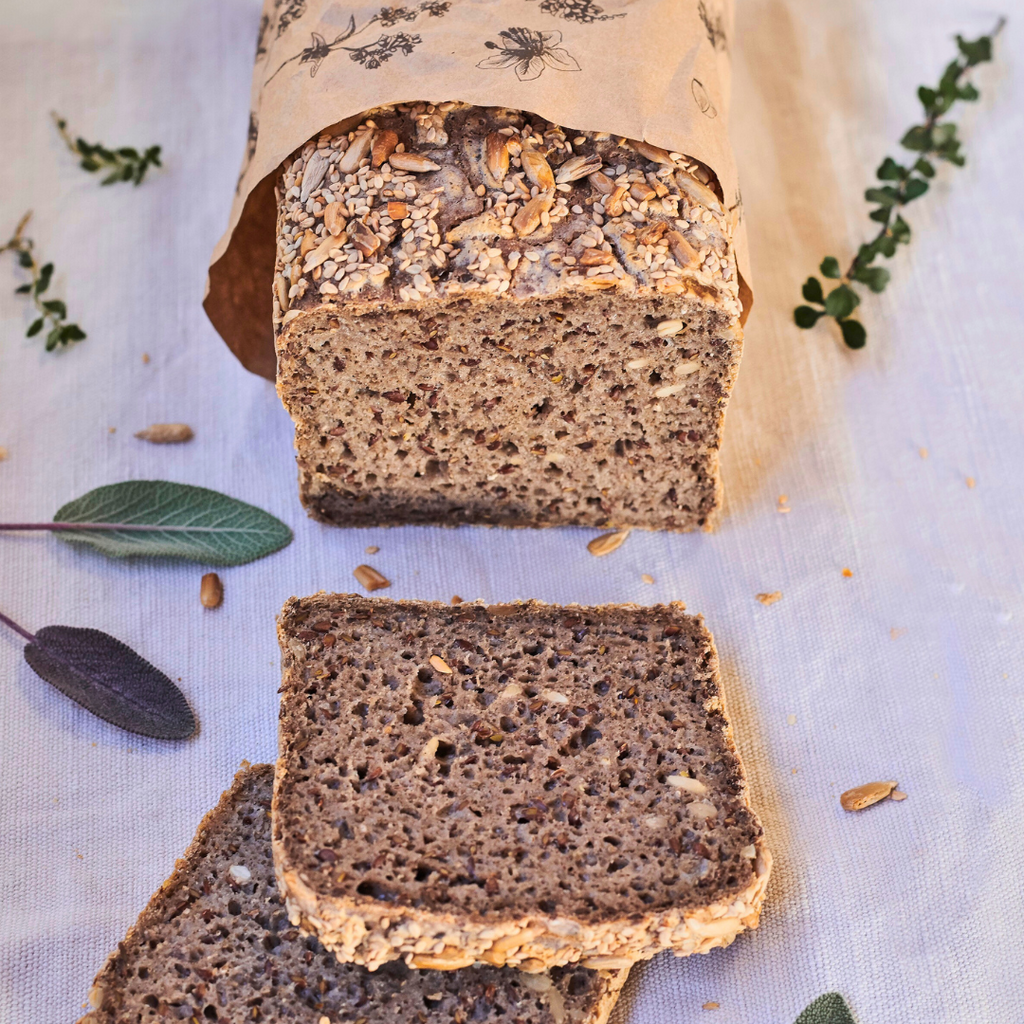 OMGoodness Sunflower & Sesame 'Wild & Free' GF Organic loaf of bread on a white background with fresh herbs sprinkled alongside. Label reads Gluten Free, Paleo & Vegan, Dairy, Egg & Refined Sugar Free