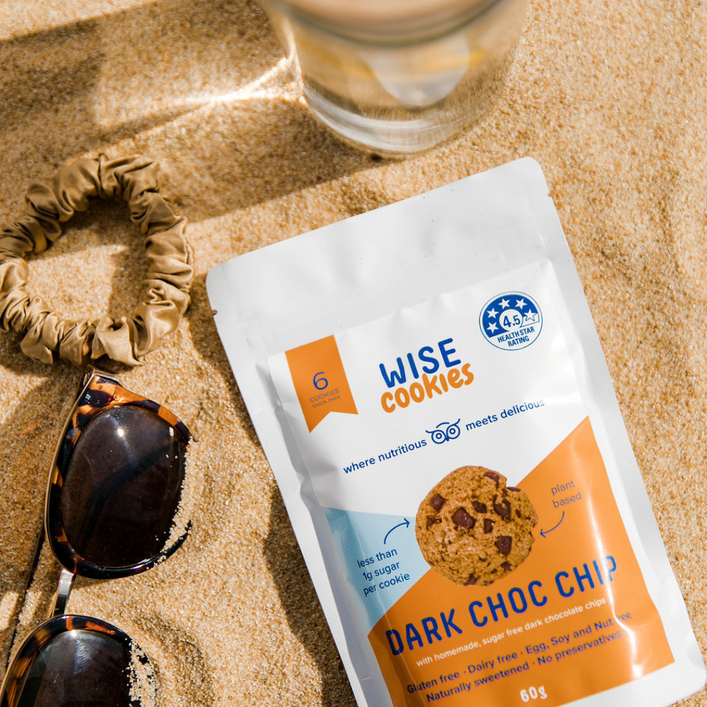 Wisefoods | A Snack Pack of Dark Choc Chip 4.5 Health Star rated, allergy-friendly and plant based Wise Cookies lying on the sand, with sunglasses, hair tie and a glass of lemon water