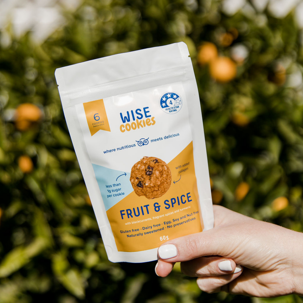 Wisefoods | Hand holding a Snack Pack of 4.0 Health Star rated, allergy-friendly Fruit & Spice Wise Cookies in front of a lemon tree out-of-focus in the background