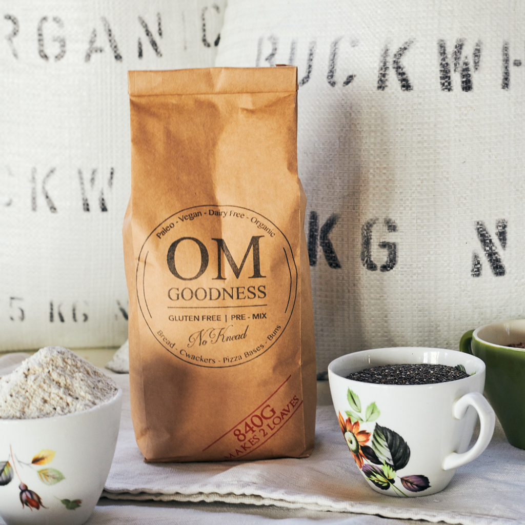 2-Loaf brown kraft bag of OMG Original Organic GF No-Knead Bread Premix standing in front of white sacks of organic buckwheat flour, with old fashioned tea cups in front filled with GF flours, chia seeds and linseeds