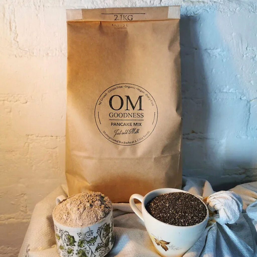 Medium-size 2.1 Kg brown kraft bag of OMG Organic GF Pancake Mix standing in front of white sacks of organic buckwheat flour, with old fashioned tea cups in front filled with GF flours, chia seeds and linseeds