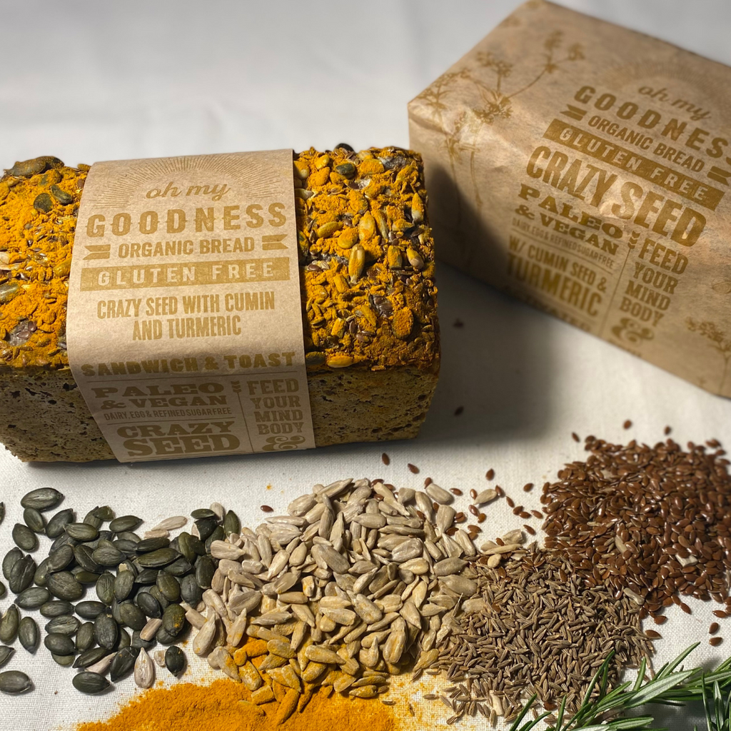 OMGoodness 'Crazy Seed' with Cumin & Turmeric GF Organic loaf of bread on a white background with pumpkin seeds, sunflower seed, cumin seeds, linseeds, turmeric powder & fresh rosemary sprinkled below. Label reads Gluten Free, Paleo & Vegan, Dairy, Egg & Refined Sugar Free