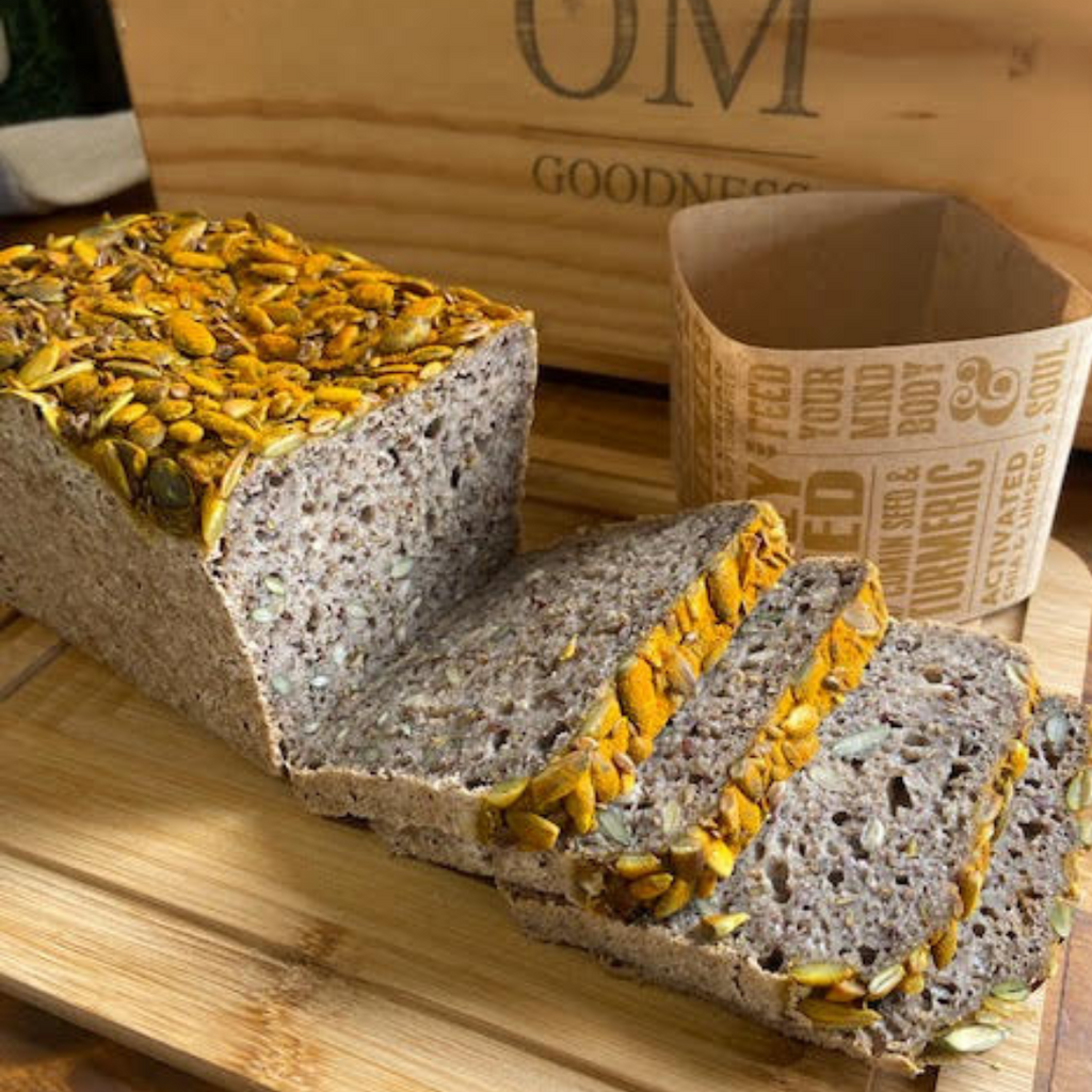 OMGoodness Sliced 'Crazy Seed' with Cumin & Turmeric GF Organic loaf of bread on a pale wooden chopping board with OMG label and inked log on a wooden box in the background. Label reads 'Feed your mind, body & soul".